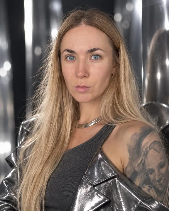 A woman with long blond hair in a silver collar and charcoal knit, a metallic jacket off one shoulder, steel bokeh behind