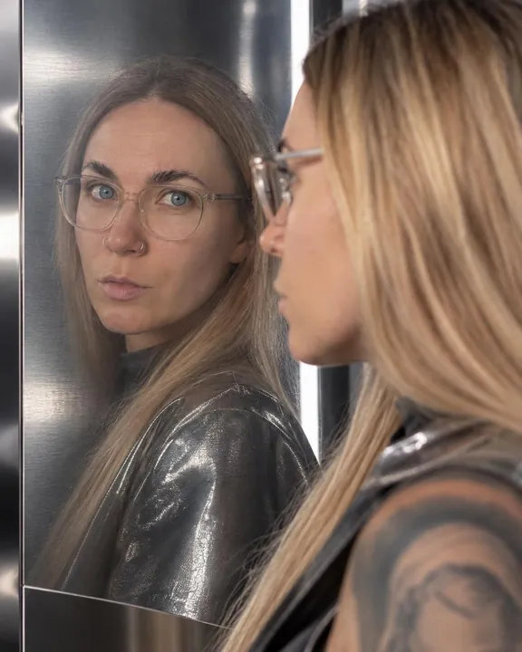 A woman with long blond hair in clear-frame glasses seen sharp in a brushed steel panel, her real profile soft in front