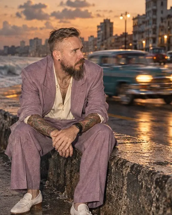 A man sits on the Malecón seawall in a faded lilac suit while a vintage car blurs past on the road