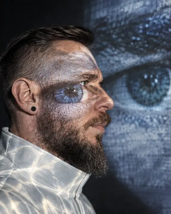 Profile of a bearded man, a huge projected fragment of his own eye falling across his cheek and the wall