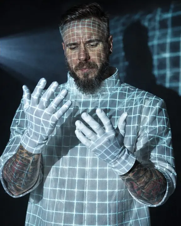 Waist-up view of a man wrapped in a fine glowing projected grid, white-gloved hands raised into the beam