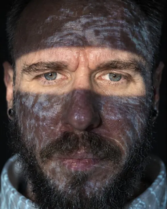 Close portrait of a bearded man with open eyes inside a band of projected light, the rest of his face textured