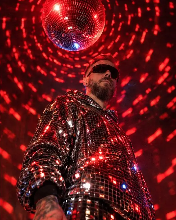Low angle on a spinning mirror ball overhead, red light arcs raining down on a man in shield sunglasses