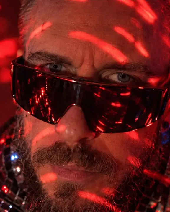 Close-up of a bearded man's sharp eyes between curving red lines of light from a disco ball