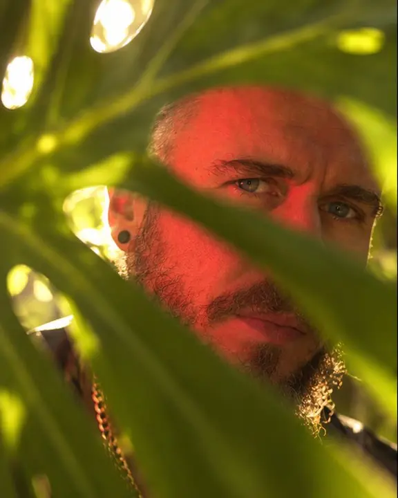 A bearded man's face seen through slits in a glowing monstera leaf, red flash light on one cheek