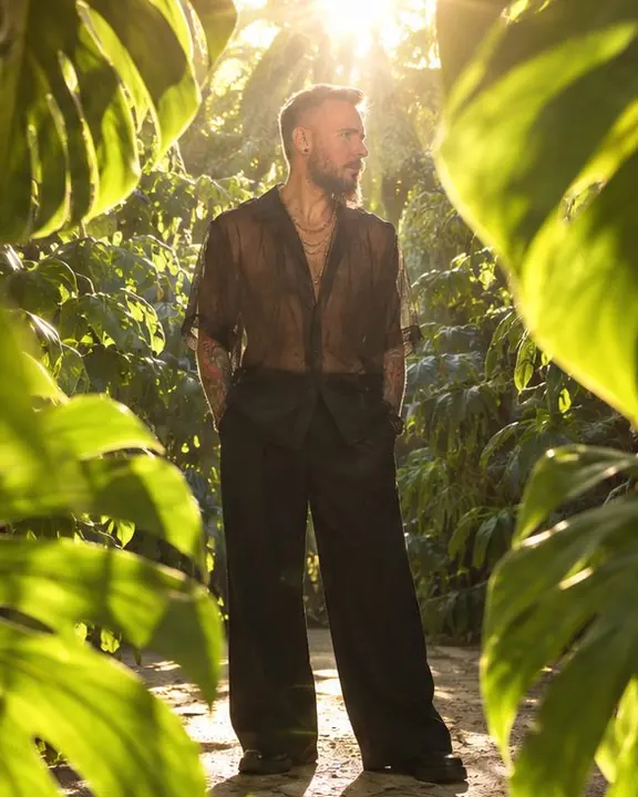 Full-length man in a sheer black organza shirt between huge backlit leaves, the sun flaring above