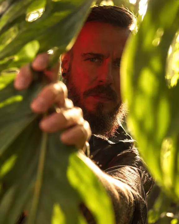 A man's hand parting large glowing tropical leaves, his bearded face in shadow behind them