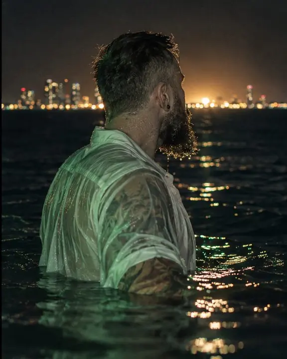 A man in a soaked white shirt turning toward the horizon, the wet fabric clinging, the sea dark around him