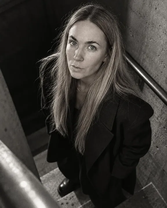 From a step above, a woman with long blond hair looks up into the lens, black coat collar, concrete wall and steel rail