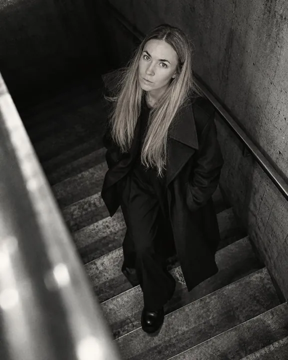 Steep top-down view of a woman with long blond hair climbing concrete stairs in a long black coat, hands in her pockets