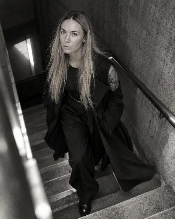 A woman with long blond hair walks up the stairwell, her long coat swinging open, wide black trousers and heavy boots