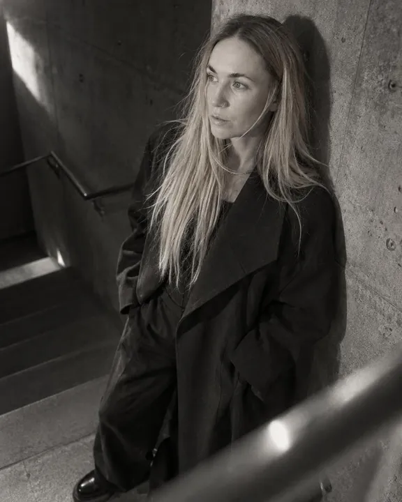 A woman with long blond hair leans on a concrete wall at the landing, looking away, hands in her coat pockets, side light
