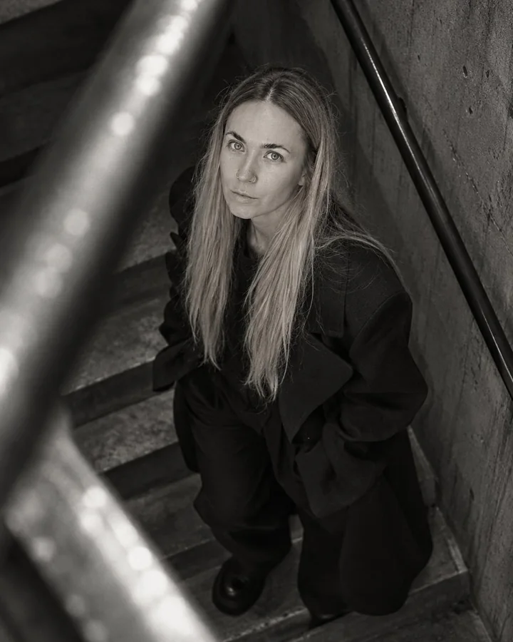 A blurred steel handrail cuts across the frame as a woman with long blond hair looks up from the stairs below