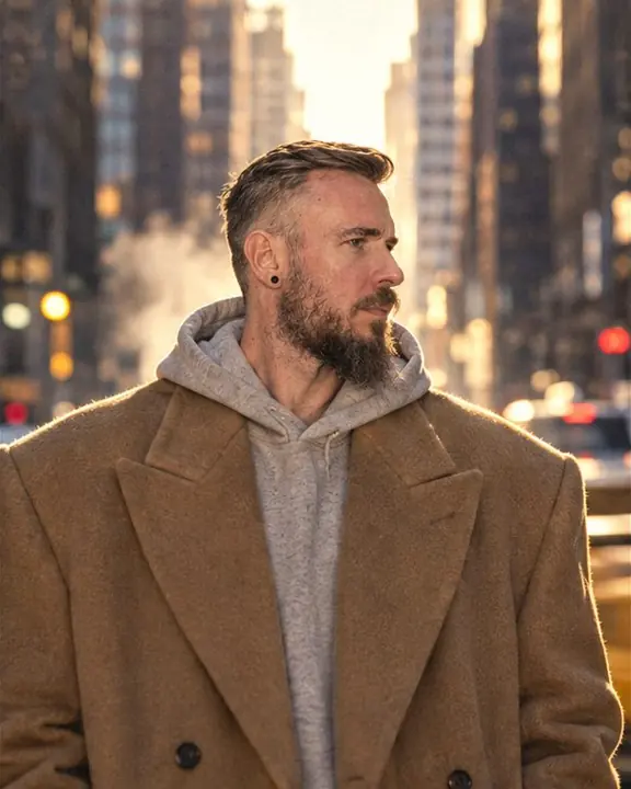 Waist-up shot of a man in a huge camel coat, skyscrapers compressed into a flat wall above his wide shoulders