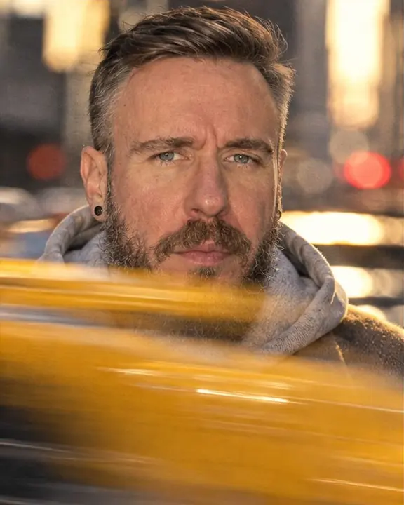 Close-up of a bearded man's face with a blurred yellow taxi streak crossing the foreground on a New York street
