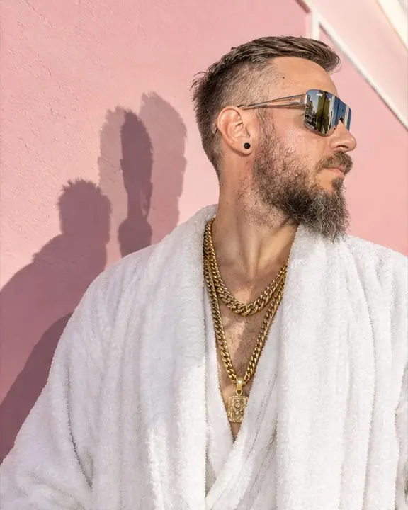 A man in white casts two hard shadows on a pink wall, one from the noon sun and one from the flash