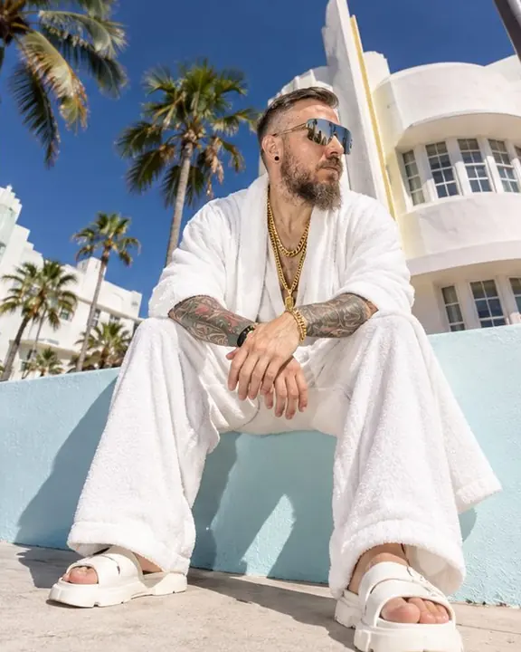 A bearded man sits on a pastel low wall in Miami with a white terry robe-coat spread around him
