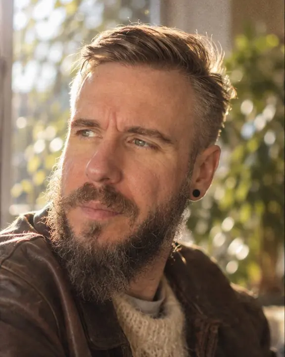 Close backlit portrait of a bearded man at a window, face three-quarters, plant leaves swirling into bokeh behind
