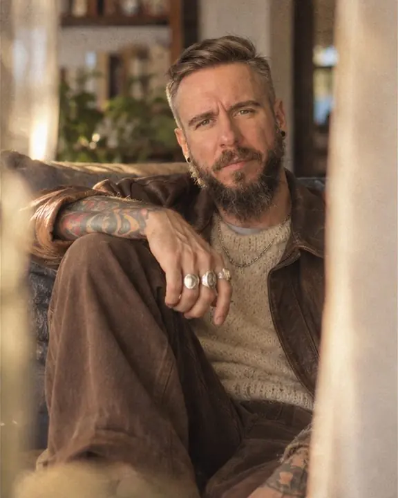 A man in a cream mohair knit sits on a worn sofa with knees drawn up, a soft curtain edge in the foreground