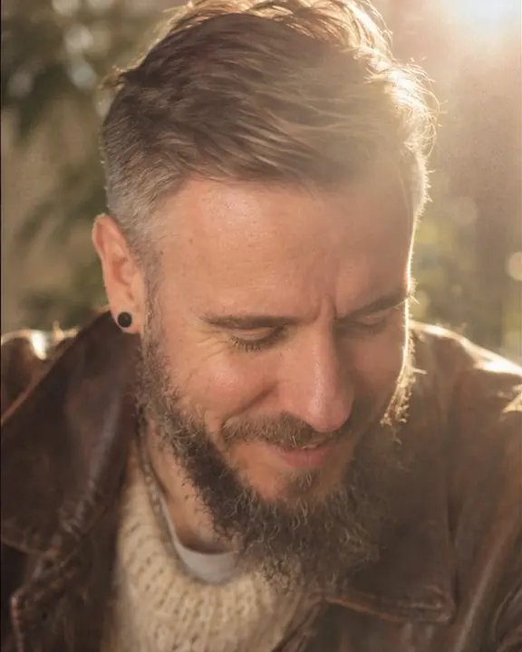 A bearded man looks down and laughs softly, head and shoulders, a warm sun flare veiling one corner of the frame