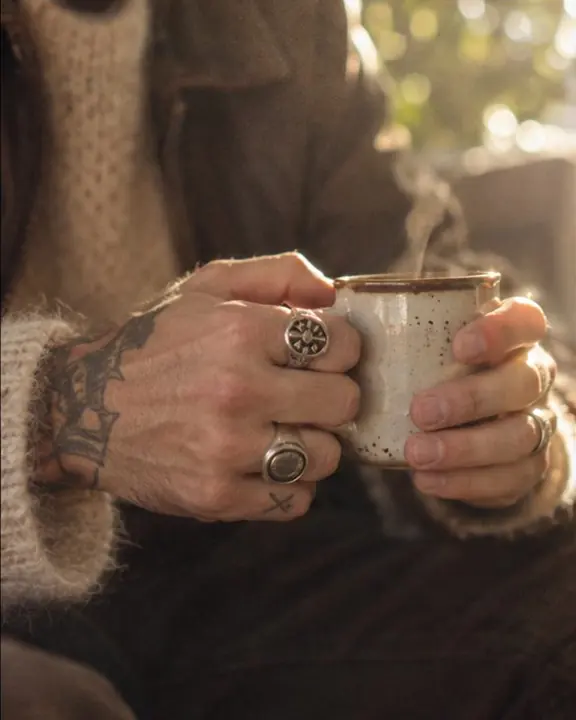 Close-up of hands with silver signet rings holding a cup in window light, cream mohair fibers glowing