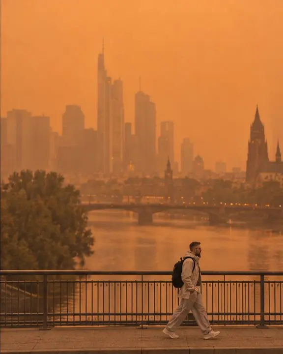 Wide shot of a man as a small figure on a bridge, the river and towers behind washed in smoky orange light