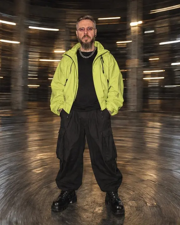 A man in an acid-lime jacket stands sharp at the center of a circular vortex of warehouse floor and light arcs