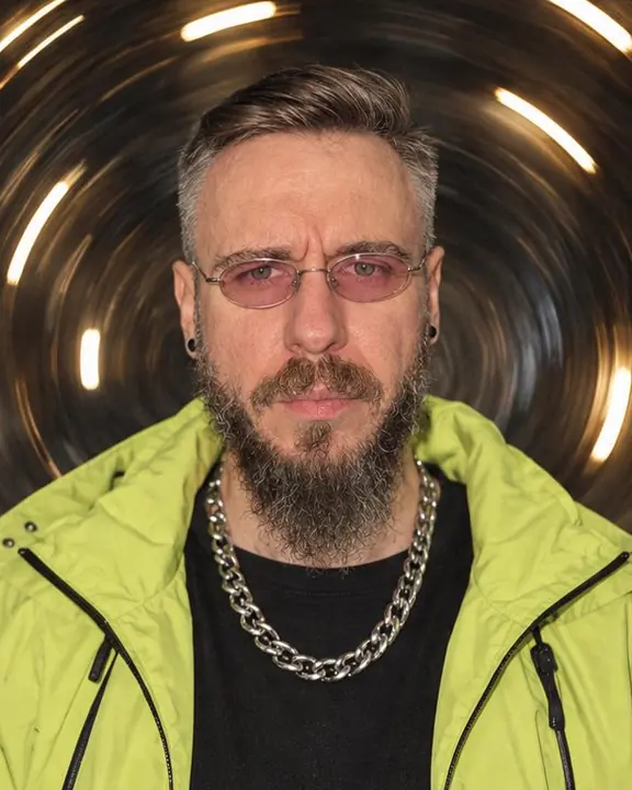 Head and shoulders of a bearded man in pink oval glasses, his face sharp inside a ring of curved light arcs