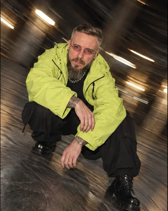 A bearded man in a lime jacket crouches and looks up, a tilted vortex of gray streaks spinning around him