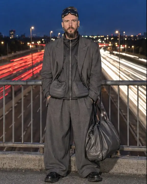 Full-length bearded man at an overpass railing at night, rivers of red tail light flowing below and behind him