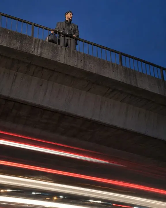 View from under a highway overpass at blue hour, a man standing above as streaks of light pass beneath