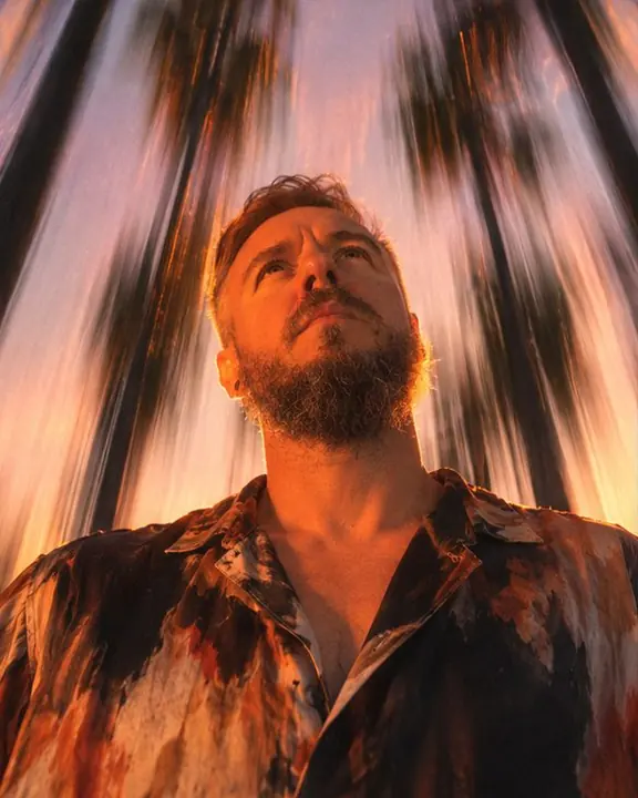 A man looking up as palm trees streak upward into a pink sunset sky in long vertical lines