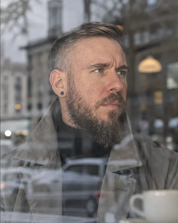 Head and shoulders of a man through a café window, street reflections across his stone-gray trench, face clear