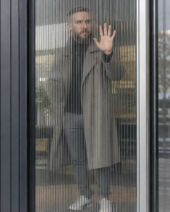 A figure behind a reeded glass door split into repeated vertical bands, one hand pressed flat and sharp on the glass