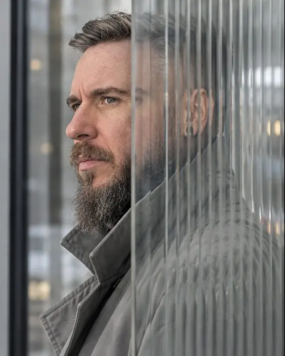 Profile of a bearded man at a glass partition edge, half his face sharp in open air, the other half repeated