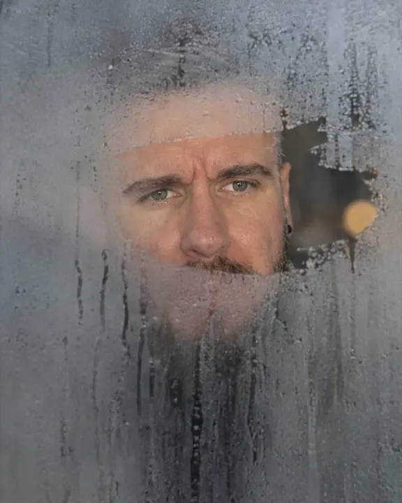 Close portrait through a steamed window, one wiped-clear patch revealing a bearded man's eyes