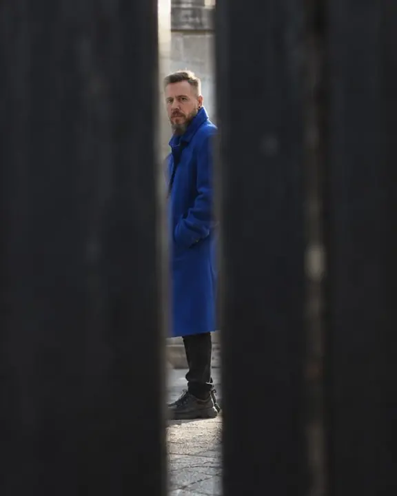 A thin slice of a man in a cobalt coat standing far away, seen through a narrow gap between dark fence planks