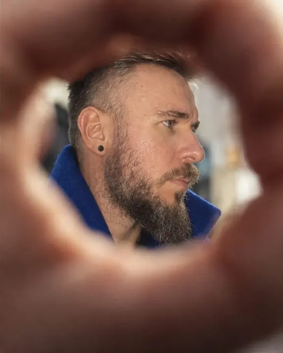 Through a ring of soft, warm fingers close to the lens, part of a bearded man's face appears in sharp focus