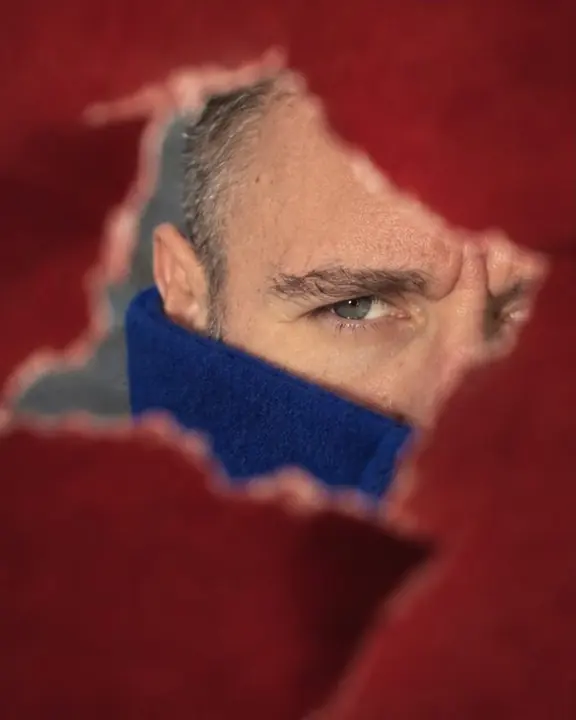 A ragged tear in red paper frames a man's eye and brow in sharp focus, with red blur all around