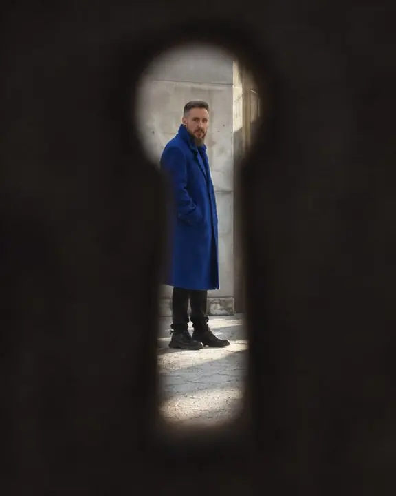 A keyhole-shaped opening in a dark door shows a man in a cobalt coat, small and full length in the light beyond