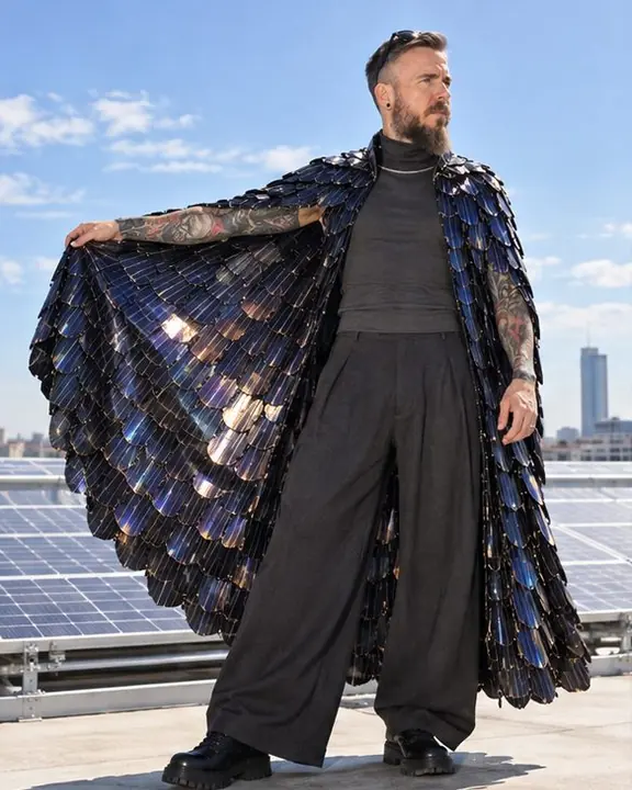 On a rooftop against open sky, a bearded man spreads a floor-length cape of iridescent solar scales with one arm