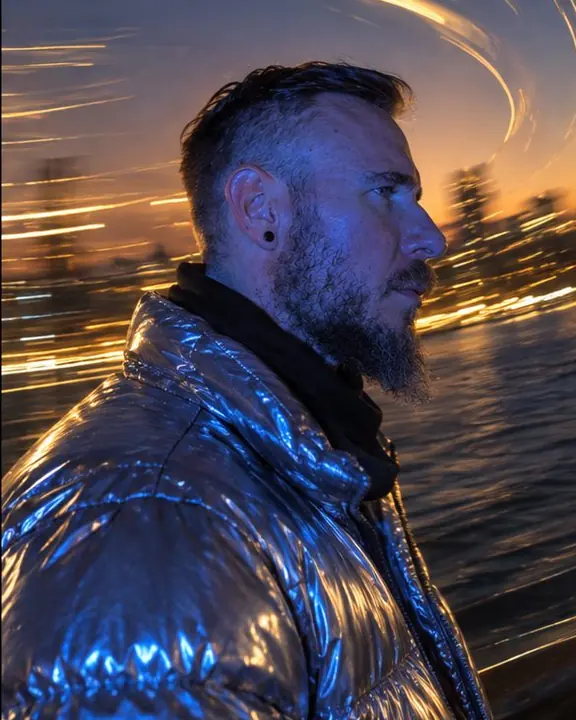 Profile of a bearded man, his chrome puffer catching blue flash, warm curved light trails behind his head