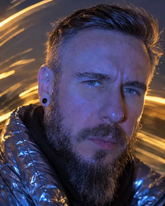 Close-up of a bearded man's eyes lit cobalt blue, warm streaks of city light across the background