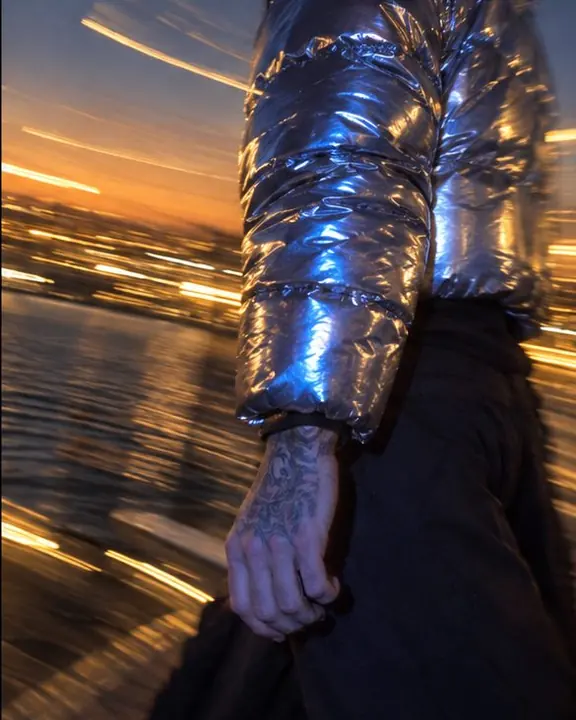 Detail of a chrome-silver puffer sleeve reflecting blue flash, golden light streaks blurred behind it