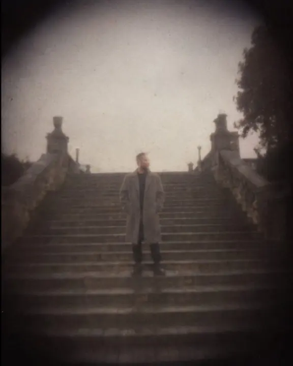 A man stands small and soft on a wide staircase with deep dark corners, faded film color and heavy vignette