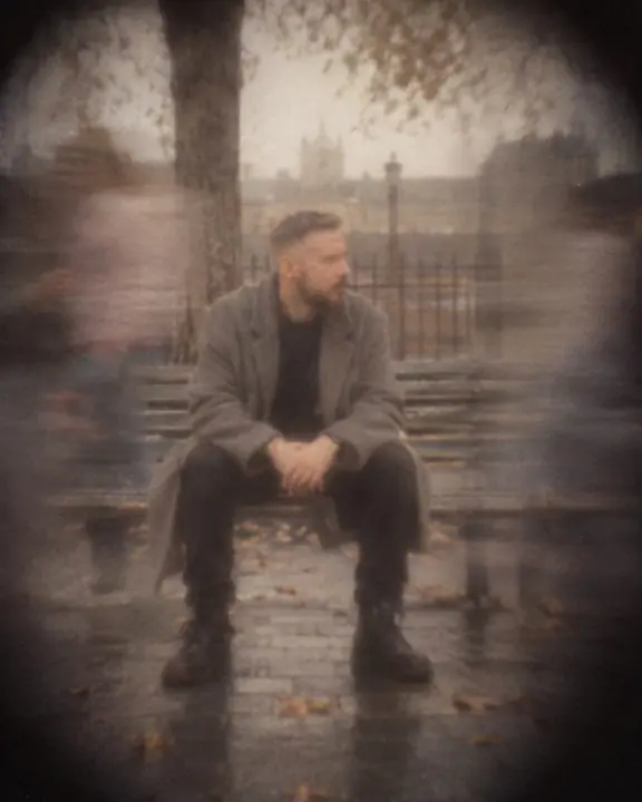 A man in a gray coat sits on a bench while translucent passersby streak past in a faded street scene