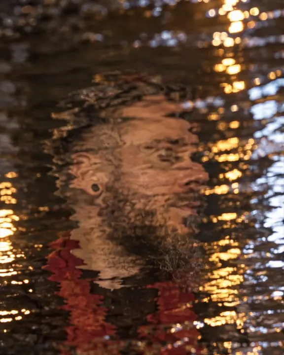A bearded man's reflected face broken into wavy strips by ripples, street lights streaking through the water
