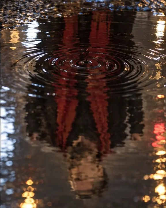 A raindrop ring spreads over the reflected chest of a man, his red scarf bleeding into the dark water