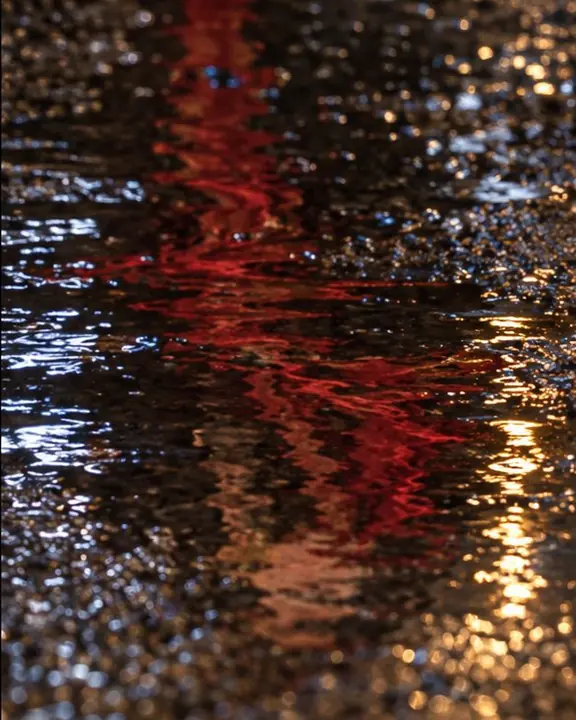 Reflection of a red scarf dissolving in rippled puddle water over sharp, textured wet asphalt