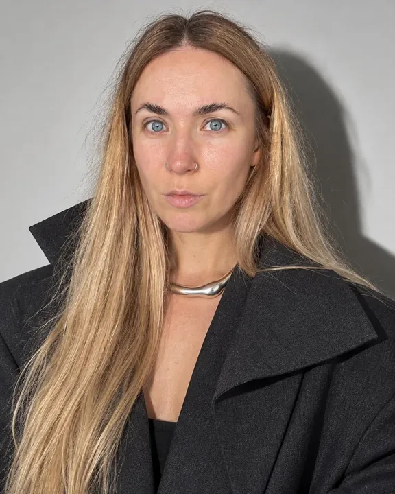 A woman with long blond hair faces the lens in a wide-collared charcoal jacket and silver collar, her shadow on the wall